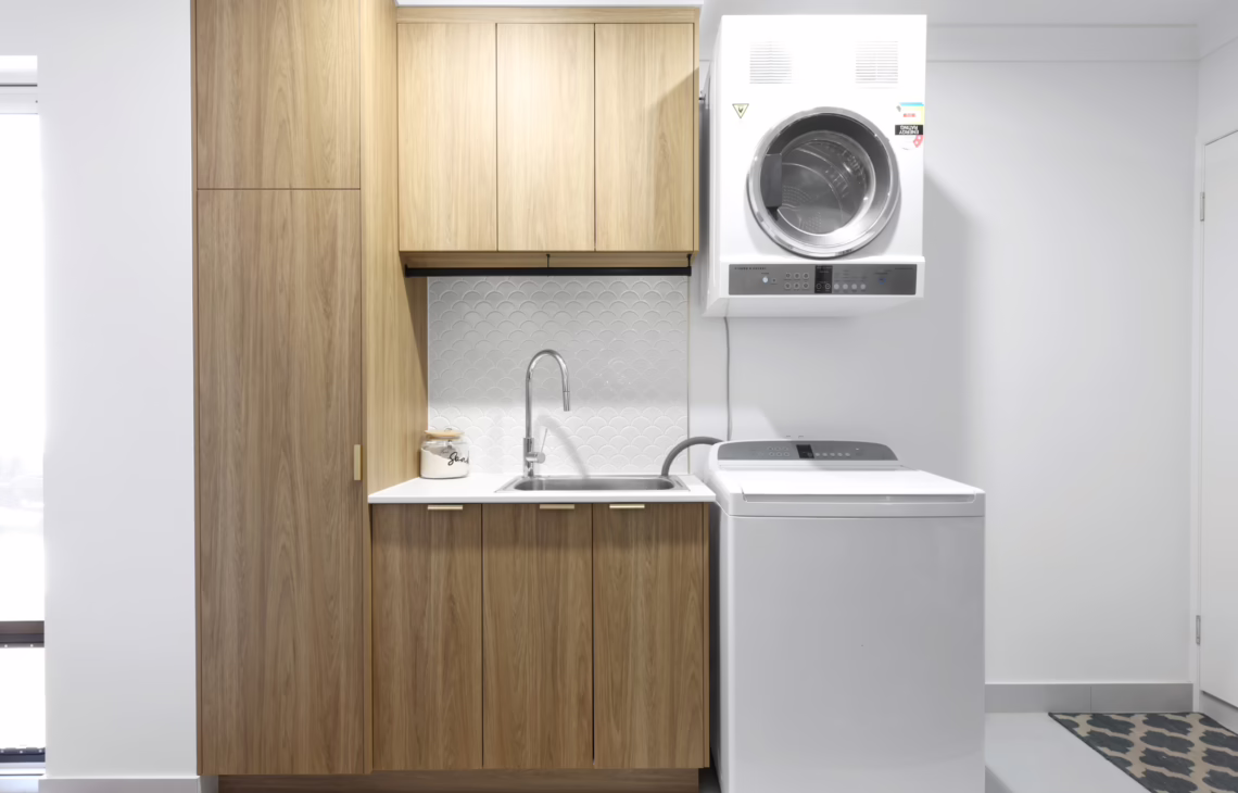 Laundry with timber cabinetry, overhead dryer, top-load washer, stainless steel sink, and white scallop splash back